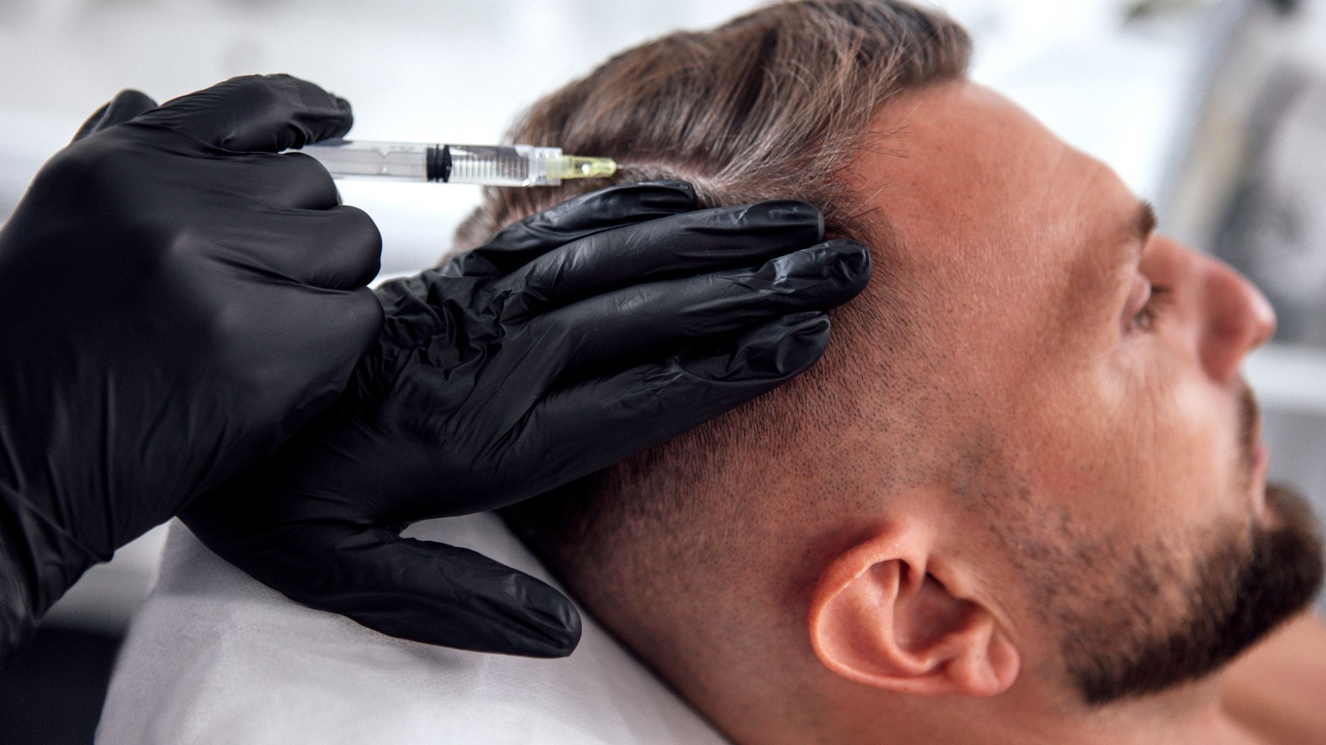 Gloved hands injecting a man’s temple with a syringe during a cosmetic procedure