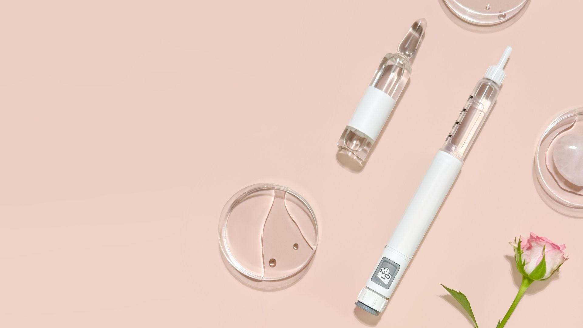 Two white cosmetic pens on a blush pink background with scattered clear glass containers and a small flower.