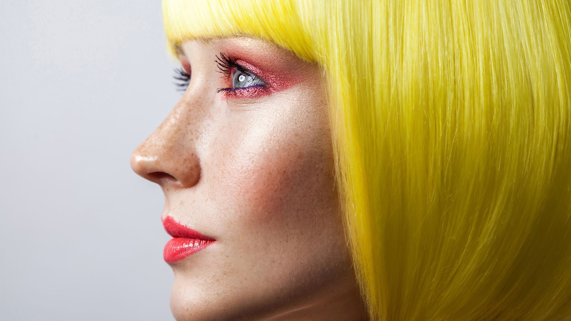Profile portrait of a person with bright yellow hair and pink eye makeup against a gray background