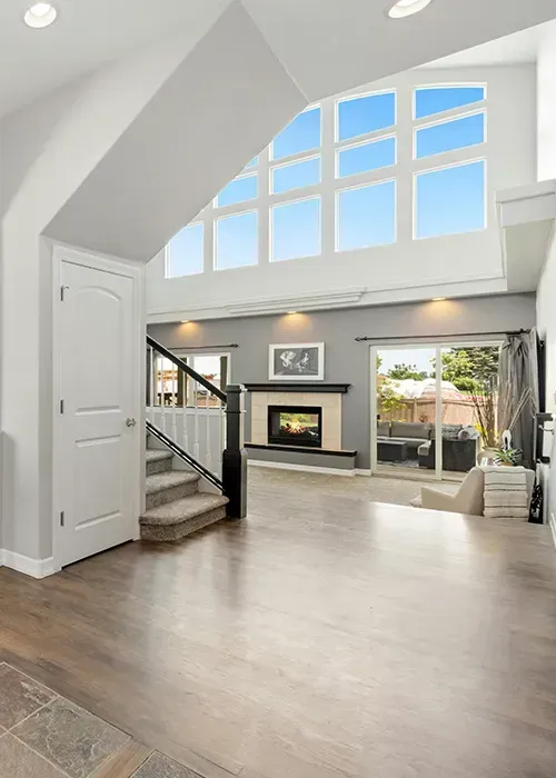 Bright modern living room with vaulted ceiling, large windows, fireplace, and open staircase