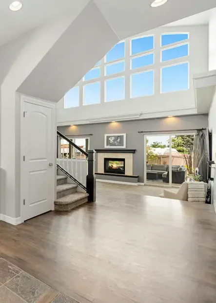 Bright modern living room with vaulted ceiling, large windows, fireplace, and open staircase