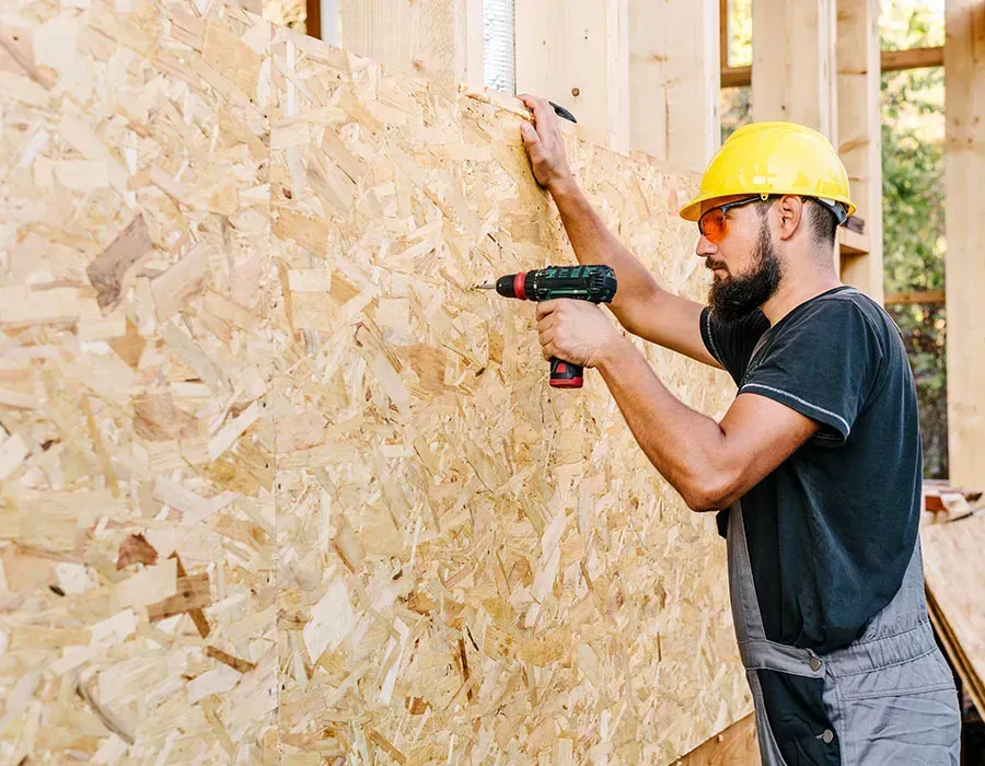 Worker in a yellow hard hat using a drill on an OSB wall at a construction site.