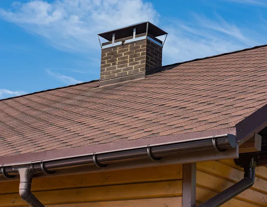 Brick chimney on a brown shingled house roof against a blue sky.