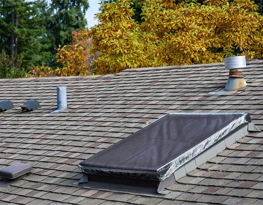 Rooftop skylight with dark cover on a shingled roof, with vents and autumn trees in the background
