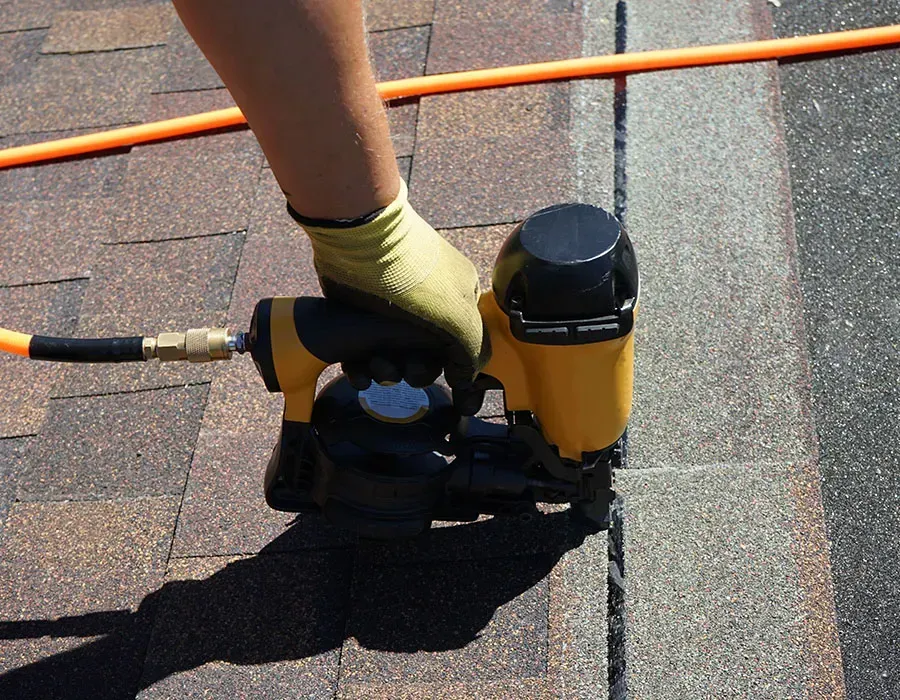 Gloved hand using a yellow and black roofing nail gun on shingles