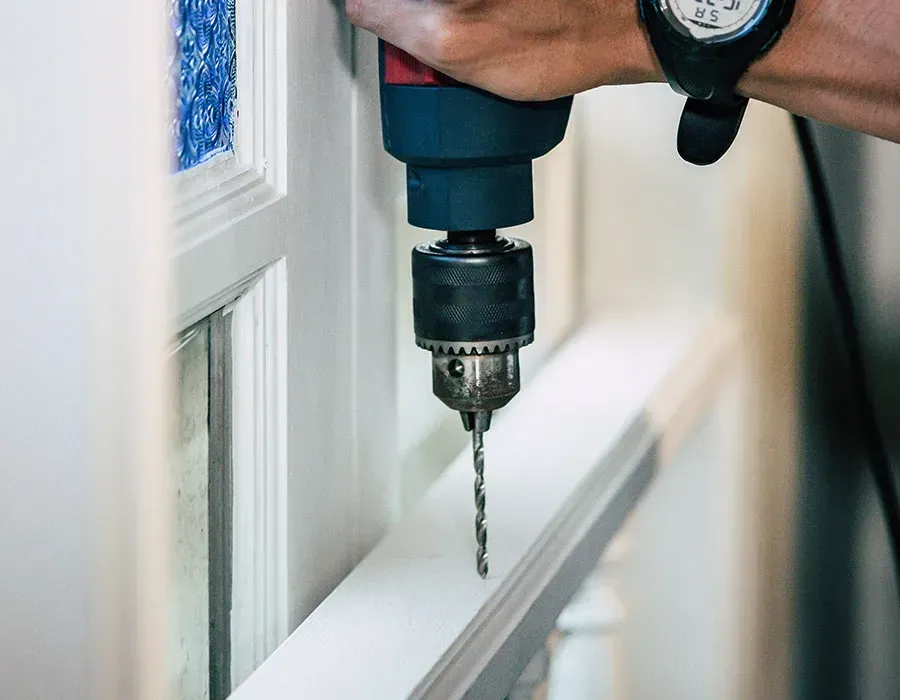 Hand holding a drill with a bit positioned near a white window frame for drilling.