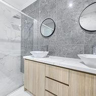 Modern bathroom with gray tile walls, glass shower, floating wood vanity, and round mirrors