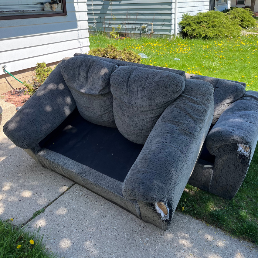 Grey, worn couch on a sidewalk next to grass. Damaged upholstery visible.