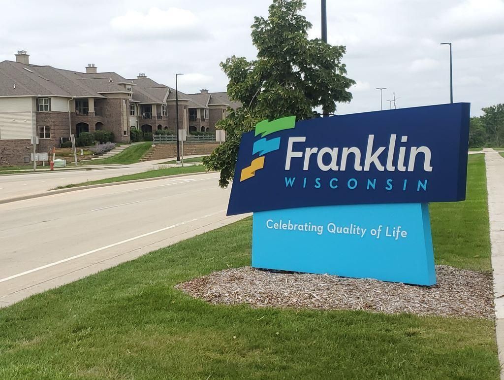 Sign for Franklin, Wisconsin, with blue, green, and gold logo; text over a blue panel. Apartment buildings in background.