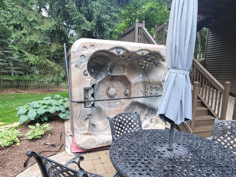 A hot tub wrapped in plastic is tilted on its side near a patio table and umbrella. Deck and foliage are in the background.