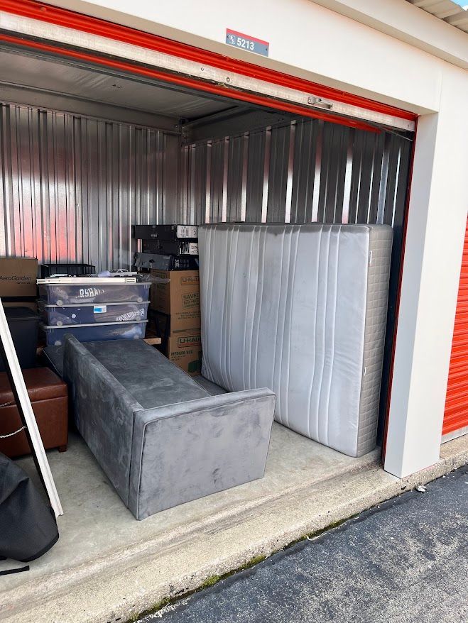 A storage unit with a mattress, bed frame, and boxes. The walls are silver. The door is open.