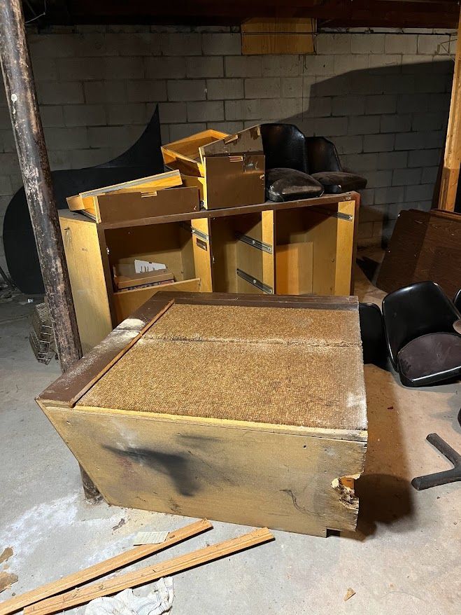 Disassembled wooden furniture in a dimly lit basement. A dusty cabinet sits in front. Other pieces and a chair are behind.