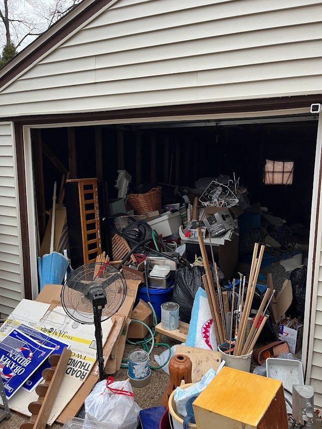Garage overflowing with clutter: bags, tools, fan, boxes, and various items. Exterior siding visible.