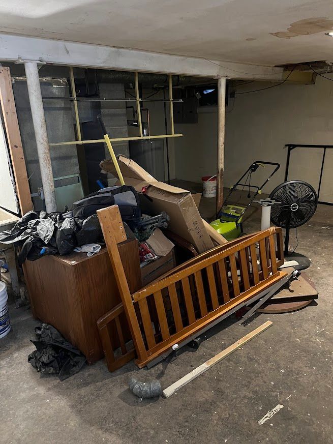 Cluttered basement with a wooden crib rail, trash bags, a lawnmower, and a fan.