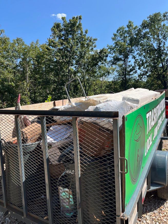 Trailer loaded with debris, including wood and foam, against a backdrop of trees and a blue sky.