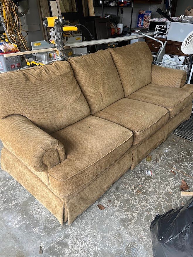 Tan three-seat couch in a garage setting with various tools and objects in the background.