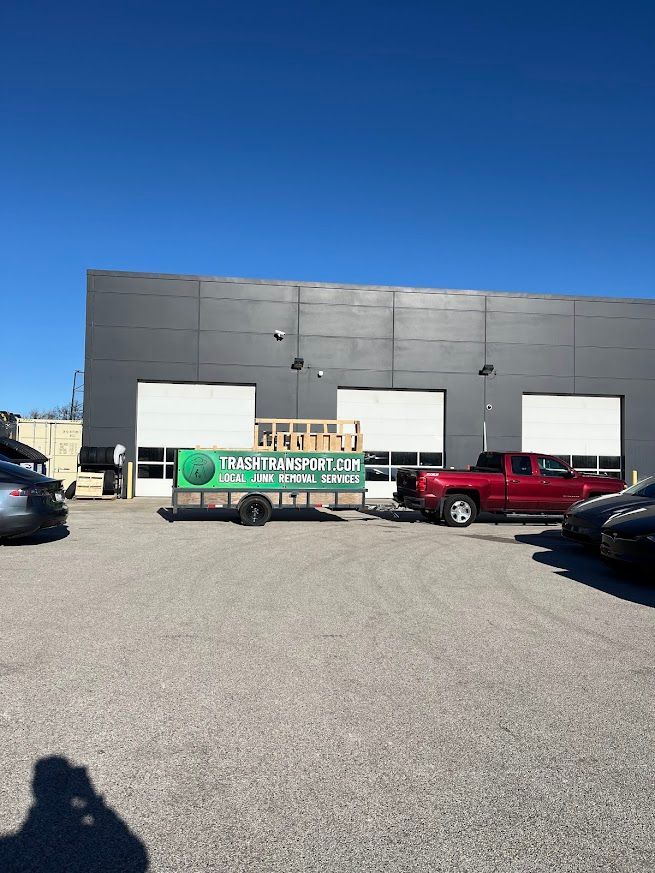 A red pickup truck pulling a trailer with a green sign in front of a gray building with loading bays.
