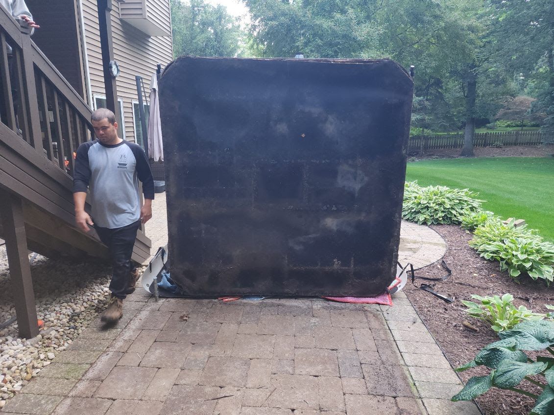 Man walking near large, black box on a brick patio. Wooden deck with stairs is on left, green lawn on right.