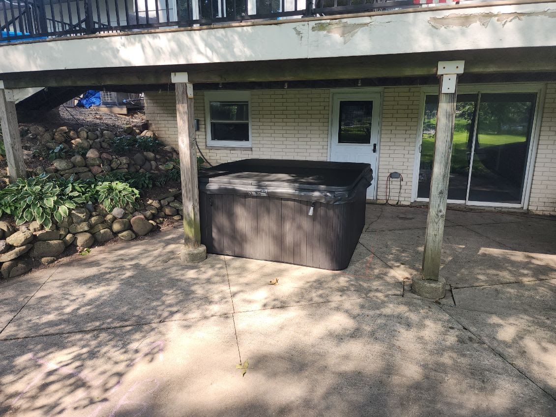 Hot tub under a deck. Concrete patio. White brick wall with door and sliding glass doors.
