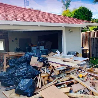 Garage with open door overflowing with debris, black bags, and wood scraps.