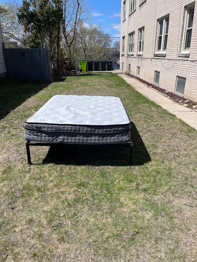Mattress on a black metal frame in a grassy backyard. A building is to the right.