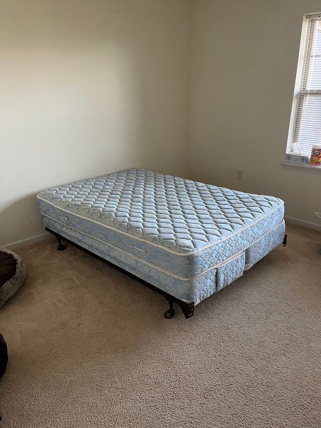 A blue mattress on a black bed frame with wheels sits in a room with beige walls and carpet.