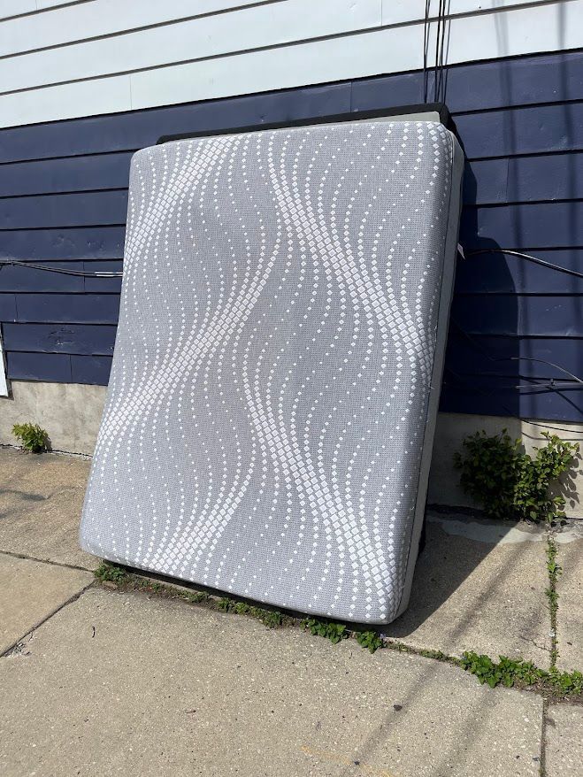 A discarded gray mattress with a white patterned top, leaning against a blue-and-white building on a concrete surface.