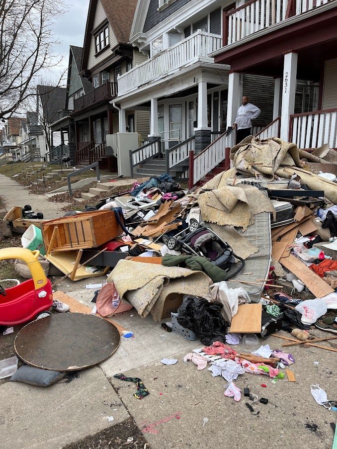 Pile of discarded household items on a sidewalk in front of a house. Person on porch.