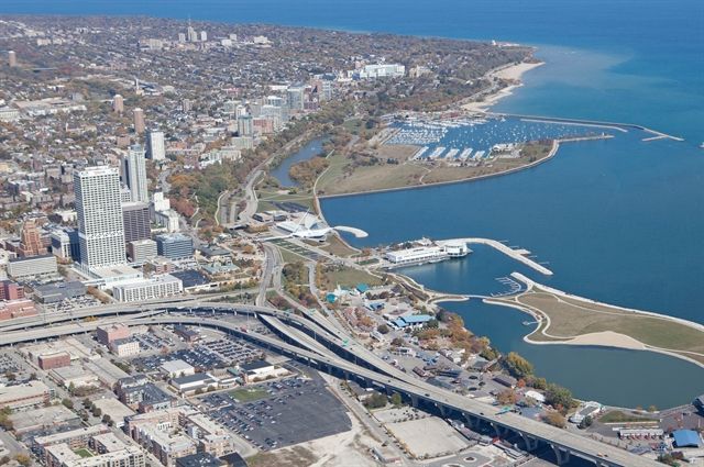 Aerial view of a city bordering a large body of water; includes buildings, highway, and a marina.