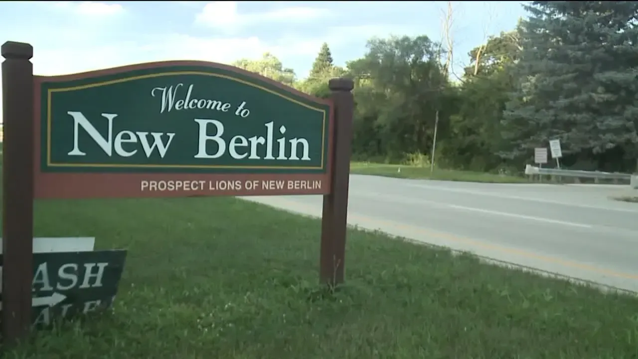 Sign welcoming visitors to New Berlin, brown posts, green grass, road in background.