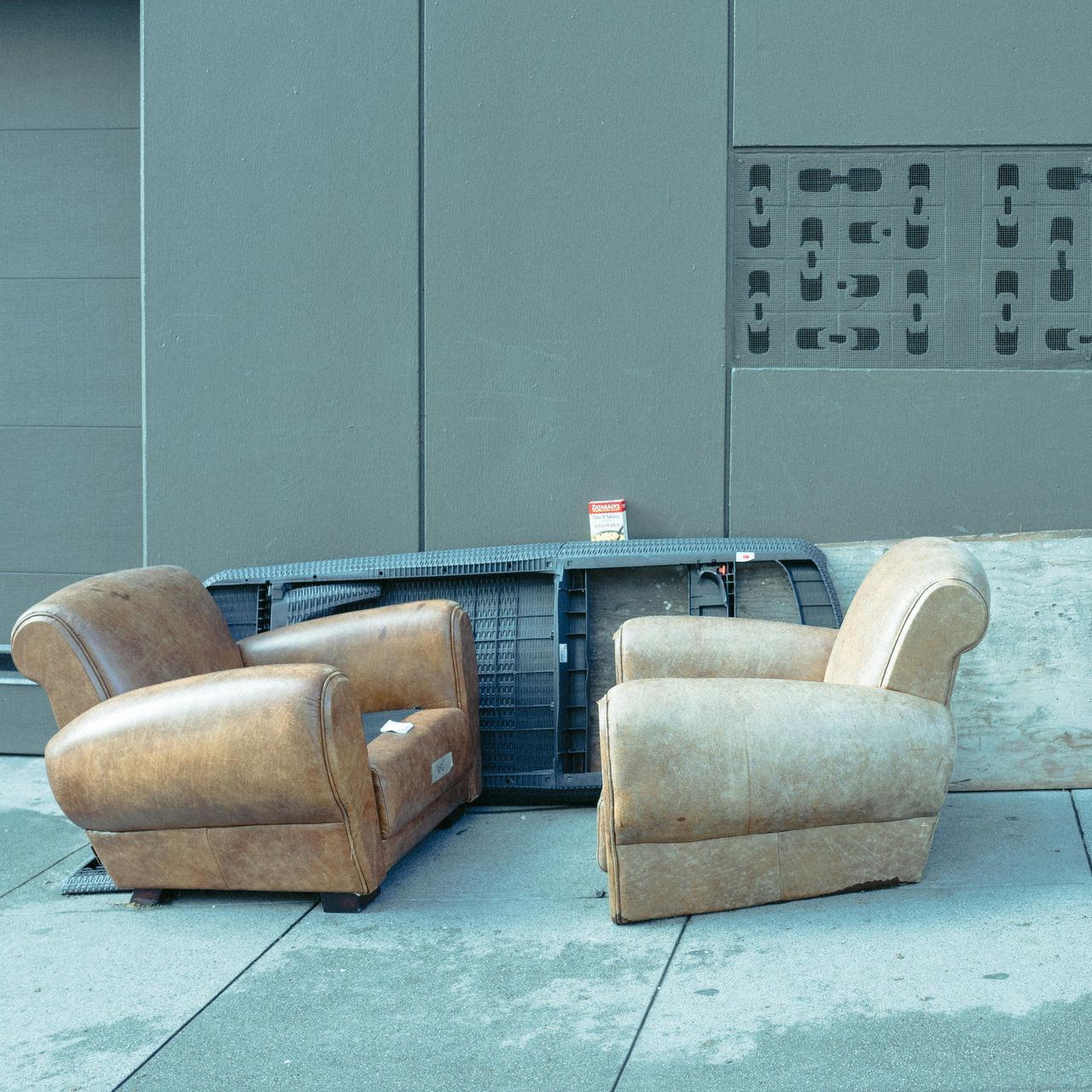 Two worn armchairs, brown and tan, outside against a gray wall and construction equipment.