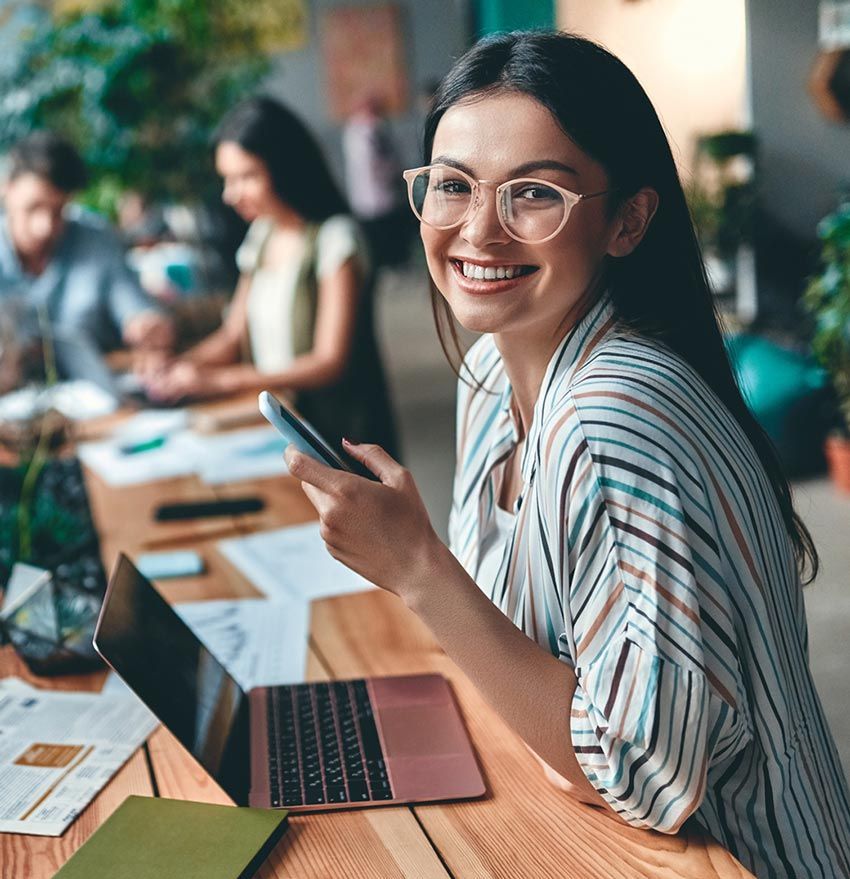 A woman is sitting at a table with a laptop and a cell phone.