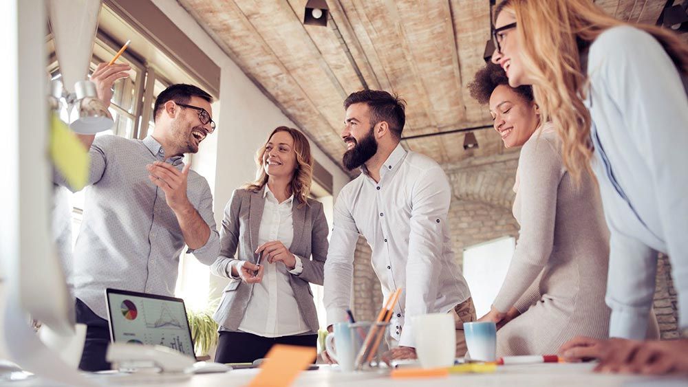 A group of people are standing around a table in an office having a meeting.