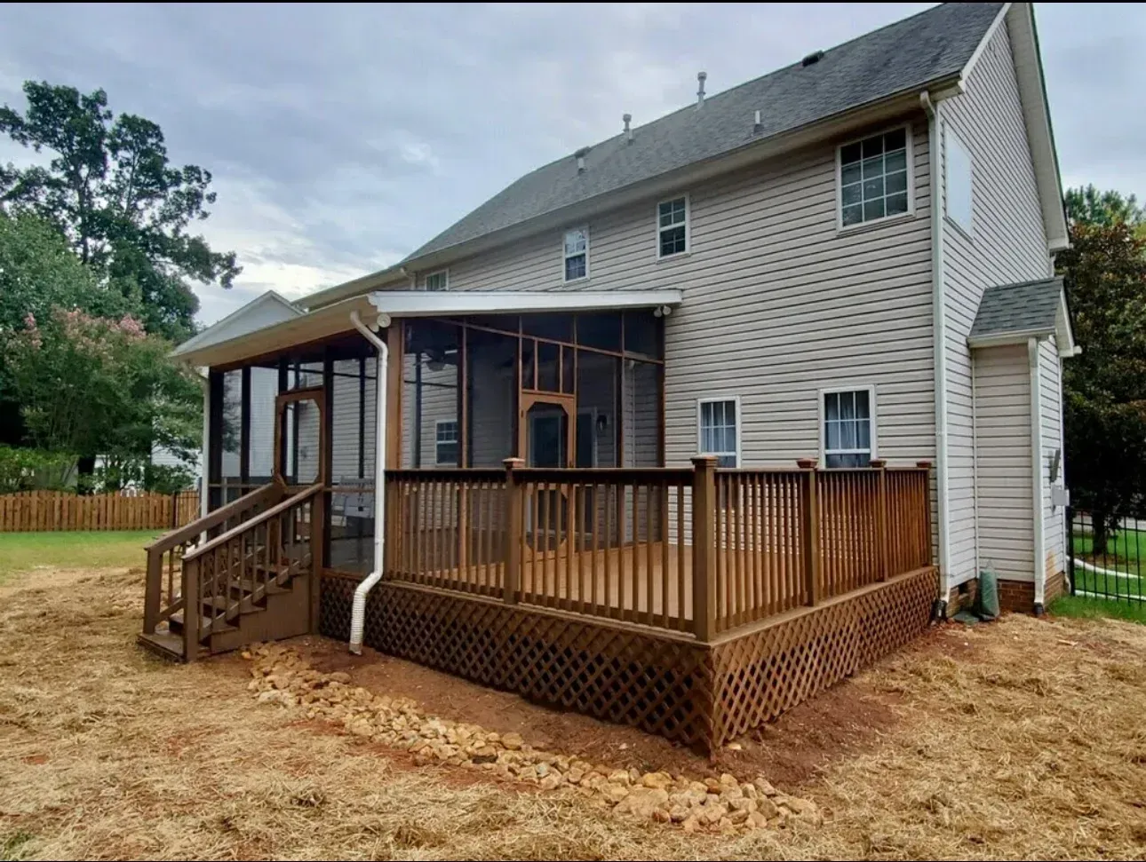 Backyard view of a two-story house with a wooden deck, screened porch, and stairs on a dirt lot