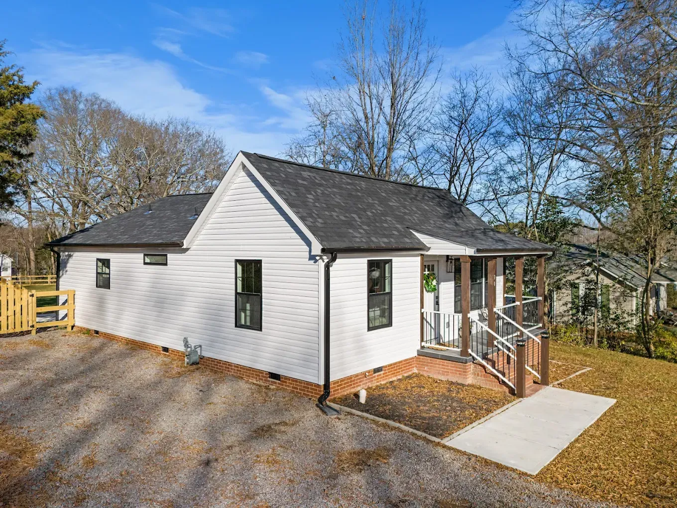 Small white house with dark roof, gravel driveway, and front porch steps surrounded by trees