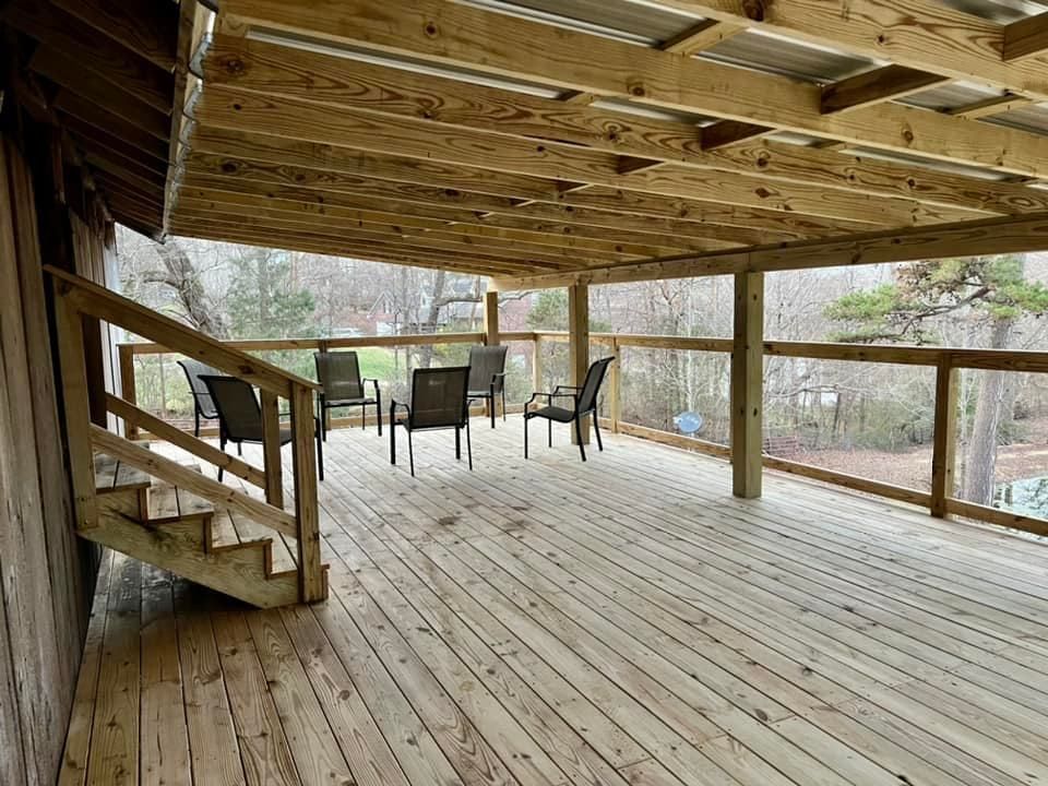 Covered wooden deck with stairs, railing, and outdoor chairs overlooking a wooded yard