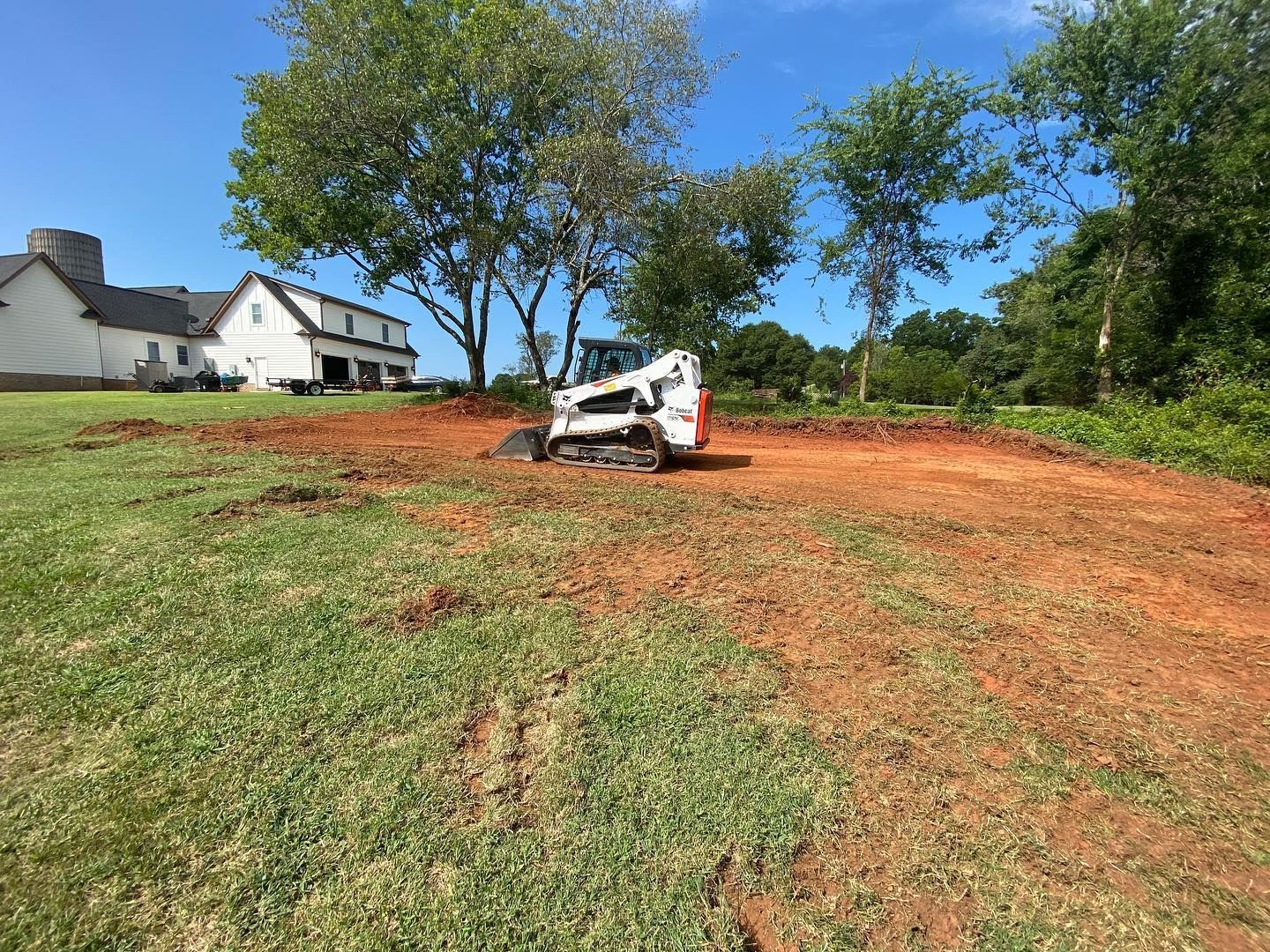 White skid-steer leveling red dirt near a house and trees on a sunny day
