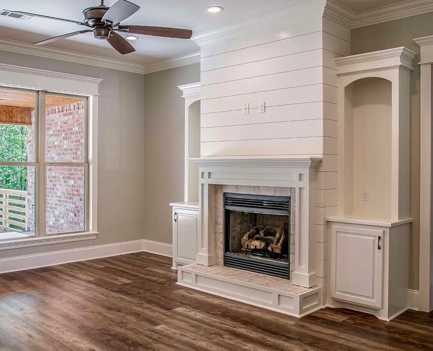 Empty living room with wood floor, white fireplace, built-in cabinets, and ceiling fan near a window