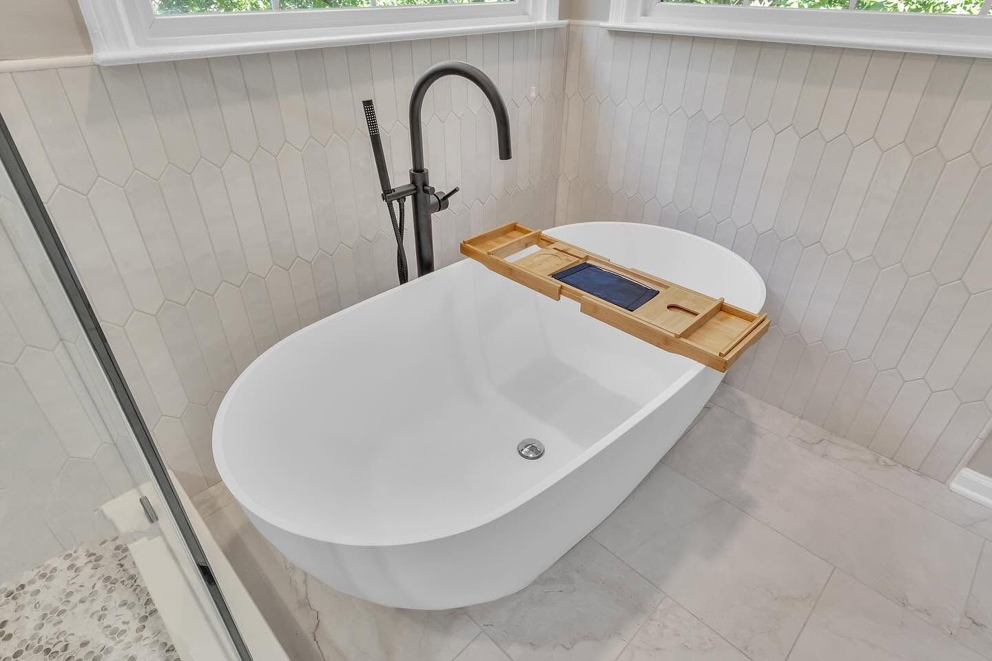 Modern bathroom with a white freestanding tub, black faucet, and wooden bath tray by a window.