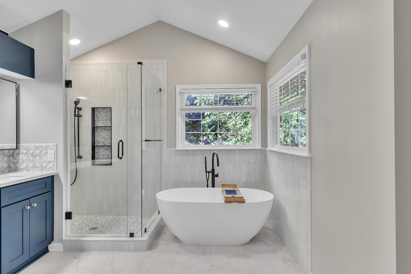 Bright modern bathroom with a freestanding tub, glass shower, blue vanity, and windows beside the tub