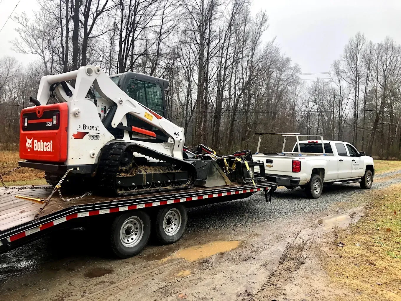 White pickup towing a tracked excavator on a flatbed trailer along a muddy road in a wooded area