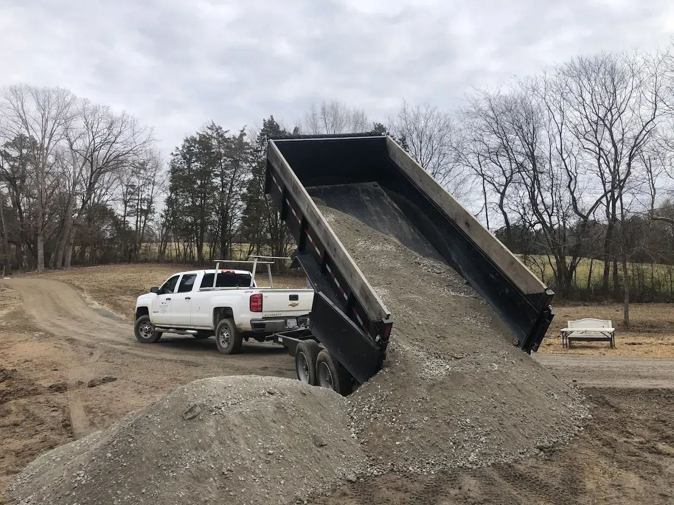 White pickup truck dumps gravel from raised bed at a construction site on a dirt road.