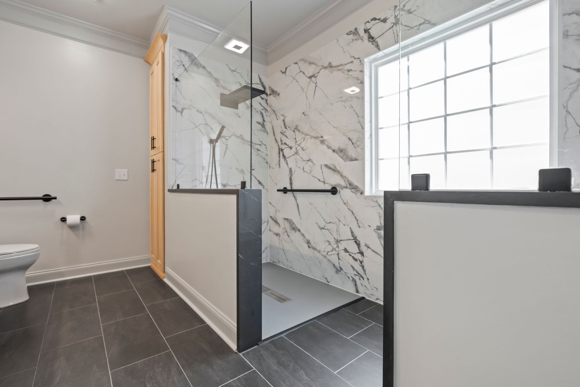 Modern bathroom with glass shower, marble-look tile, dark floor, and frosted window light