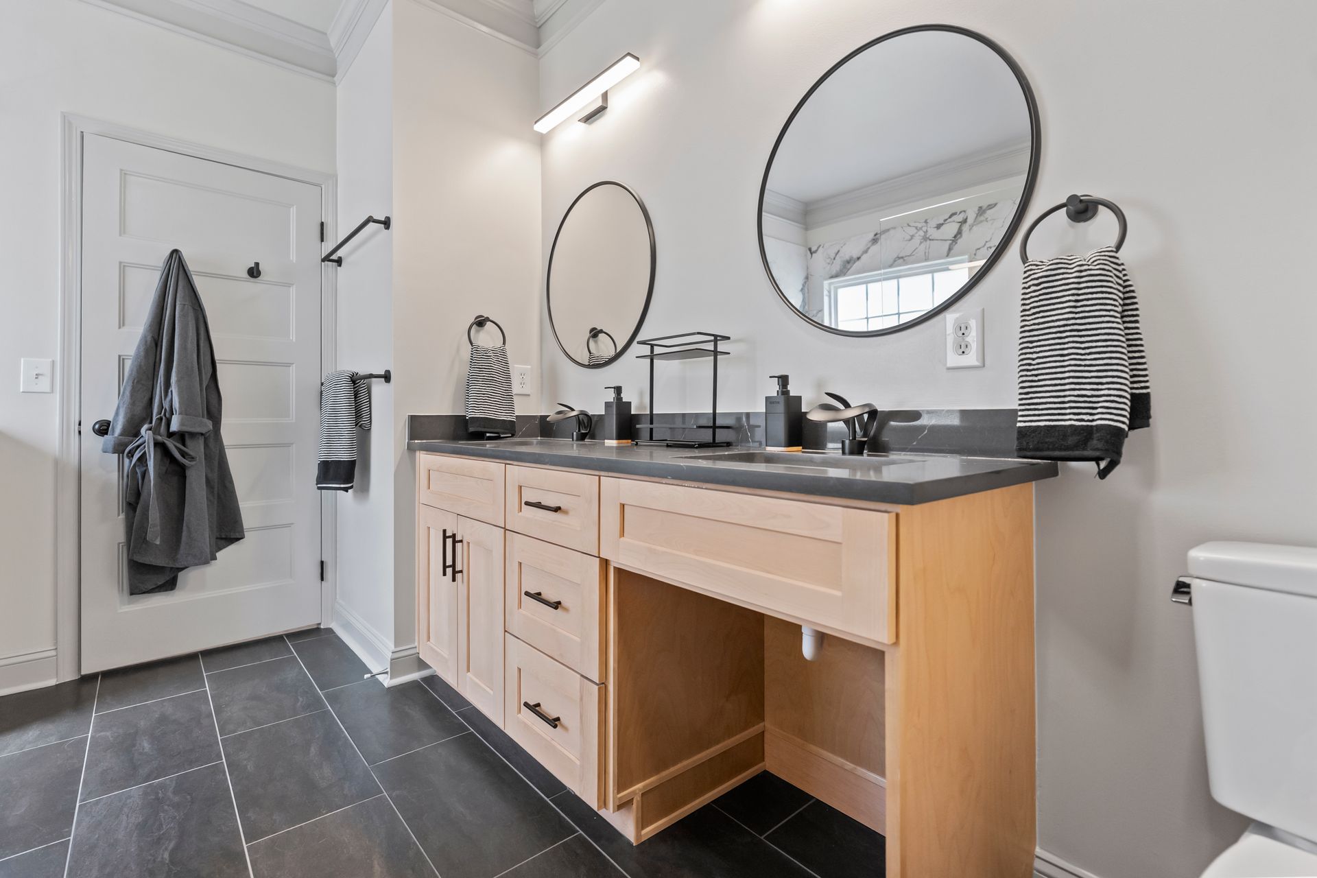 Modern bathroom with double vanity, round mirrors, gray tile floor, and towels hanging by the door