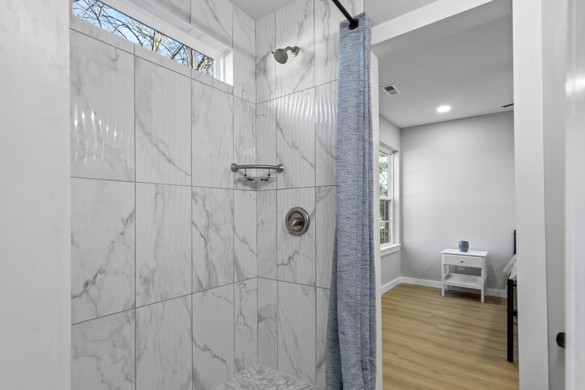 Bright tiled bathroom with a glass shower, blue curtain, and wood flooring leading to a small room beyond