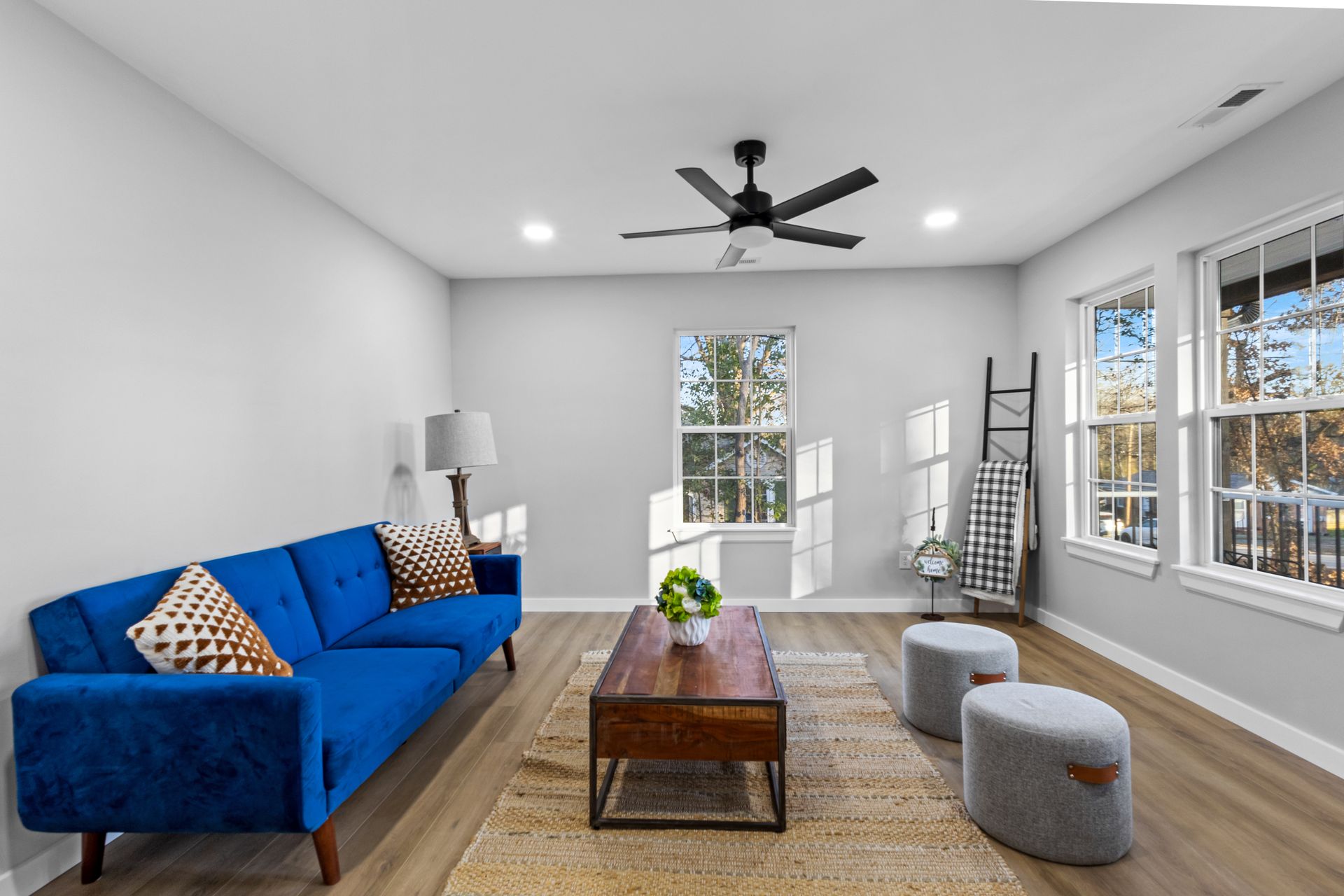 Bright living room with blue sofa, wooden coffee table, rug, gray stools, and large windows.