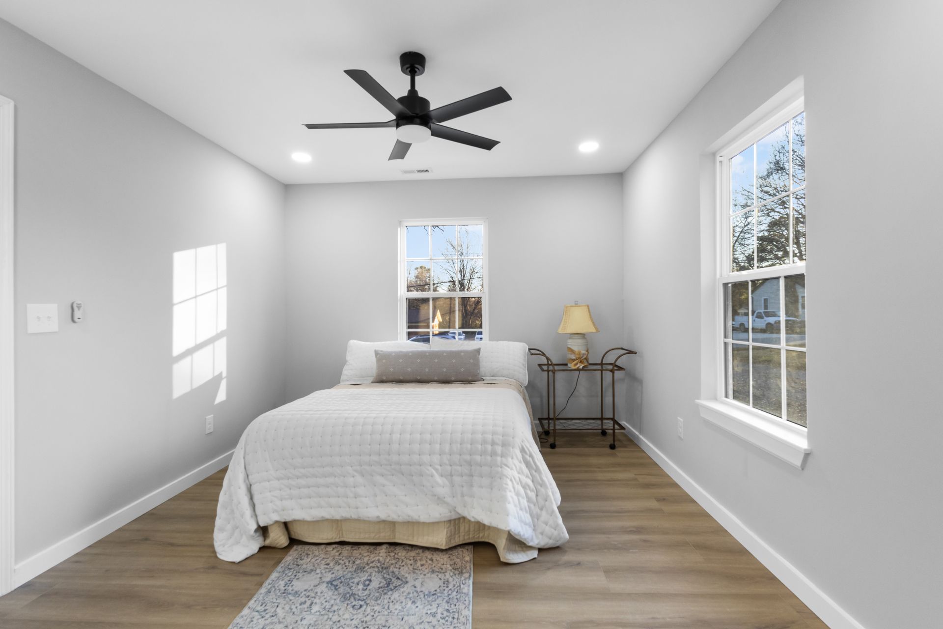 Bright bedroom with a bed, two windows, gray walls, wood floor, and a ceiling fan.