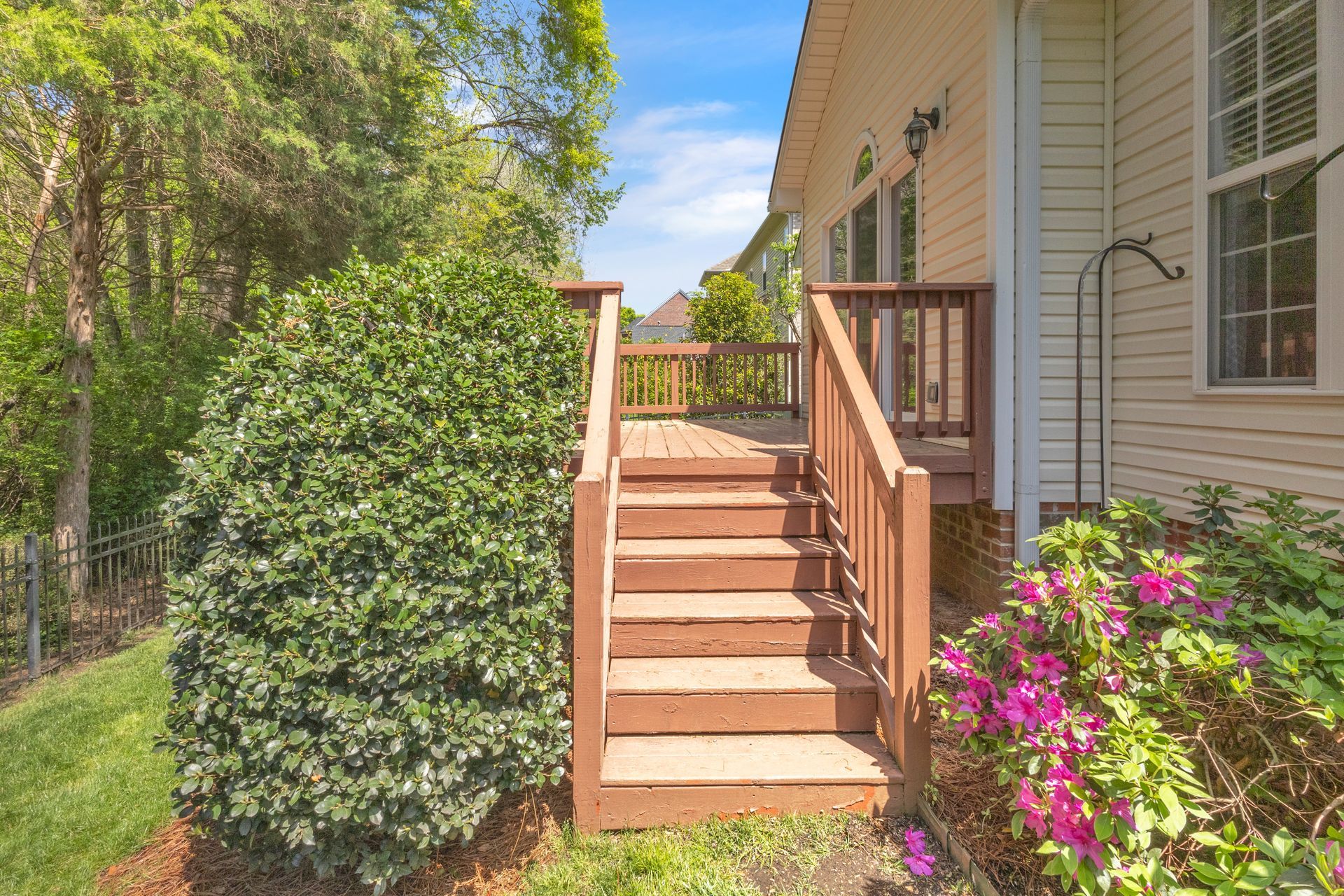 Front steps to a house with wooden railings, surrounded by green hedges and pink flowers.