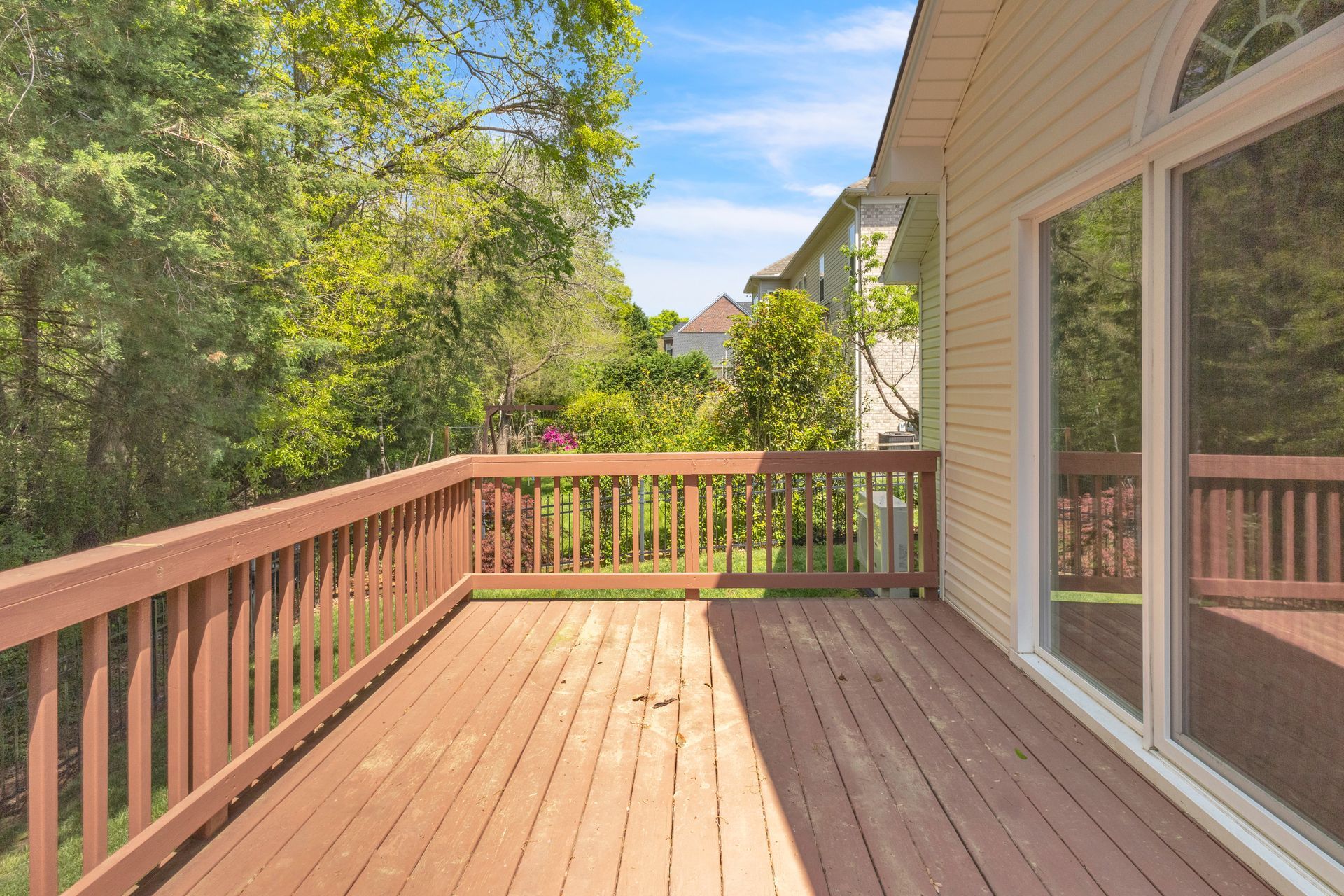 Wooden backyard deck with brown railing beside a house, overlooking trees and a sunny yard