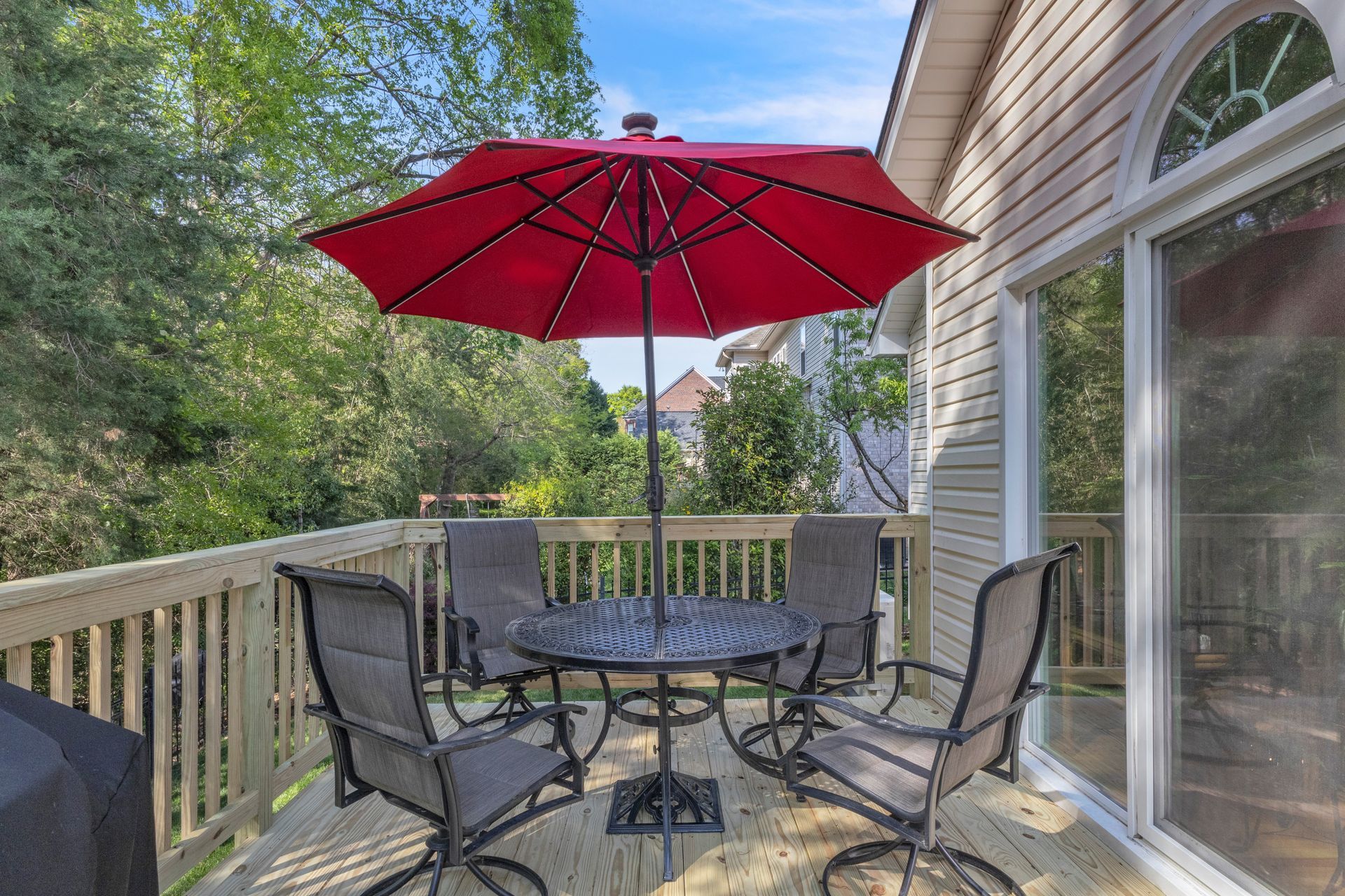 Covered deck with outdoor table, four chairs, and a red umbrella beside a house.